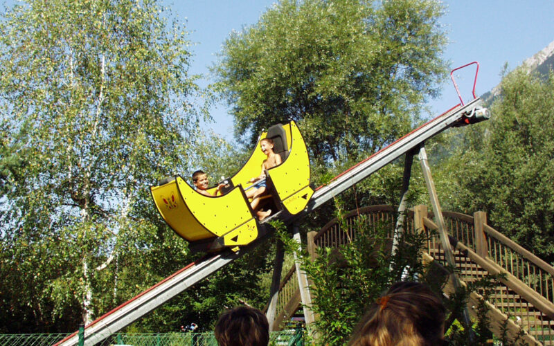 Zwei Personen fahren auf einer kleinen gelben Achterbahn eine abschüssige Strecke im Freien hinunter, wobei im Vordergrund Bäume und Zuschauer zu sehen sind.