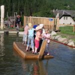 Eine Gruppe kleiner Kinder steht auf einem Holzfloß, das ein Seil hält, und überquert einen flachen Teich in einem Spielbereich im Freien mit Bäumen und Gebäuden im Hintergrund.