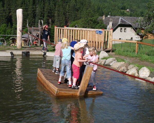 Eine Gruppe kleiner Kinder steht auf einem Holzfloß, das ein Seil hält, und überquert einen flachen Teich in einem Spielbereich im Freien mit Bäumen und Gebäuden im Hintergrund.