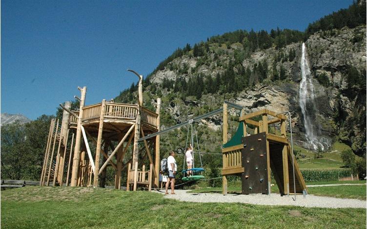 Ein hölzerner Spielplatz mit Schaukeln und Klettergerüsten liegt auf einer Wiese in der Nähe eines Wasserfalls und bewaldeter Berge unter einem klaren blauen Himmel.