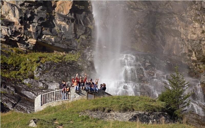 Eine Gruppe von Menschen steht auf einer steinernen Aussichtsplattform und winkt in die Kamera, während im Hintergrund ein Wasserfall und felsige Klippen zu sehen sind.