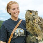 Eine Frau, die ein dunkles T-Shirt mit einem Logo trägt, steht im Freien, lächelt und hält eine große Eule in ihrer behandschuhten Hand. Im Hintergrund sind bewaldete Hügel und ein bewölkter Himmel zu sehen.