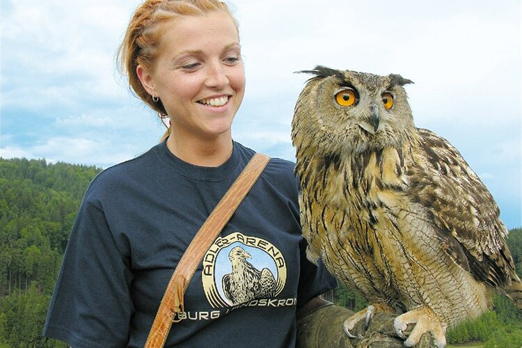 Eine Frau, die ein dunkles T-Shirt mit einem Logo trägt, steht im Freien, lächelt und hält eine große Eule in ihrer behandschuhten Hand. Im Hintergrund sind bewaldete Hügel und ein bewölkter Himmel zu sehen.