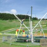 Kinder benutzen ein Bungee-Trampolin in einem Freiluftpark mit grasbewachsenen Hügeln, einem Wald und Menschen, die sich in der Nähe entspannen.