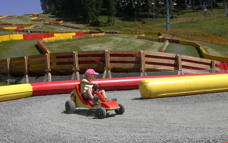 Ein Kind mit einer rosafarbenen Mütze fährt ein kleines Gokart auf einer Schotterpiste mit kurvenreichen Hindernissen und grasbewachsenen Hügeln im Hintergrund.