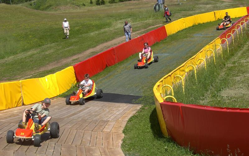 Vier Personen fahren Gokarts auf einer eingezäunten, mit roten und gelben Absperrungen versehenen Strecke, während einige Zuschauer auf einer Wiese in der Nähe zusehen.