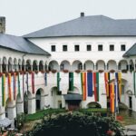 Ein Innenhof eines historischen Gebäudes mit gewölbten Gängen, roten Blumen und verschiedenen bunten vertikalen Fahnen, die von der oberen Ebene herabhängen.