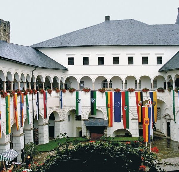 Ein Innenhof eines historischen Gebäudes mit gewölbten Gängen, roten Blumen und verschiedenen bunten vertikalen Fahnen, die von der oberen Ebene herabhängen.