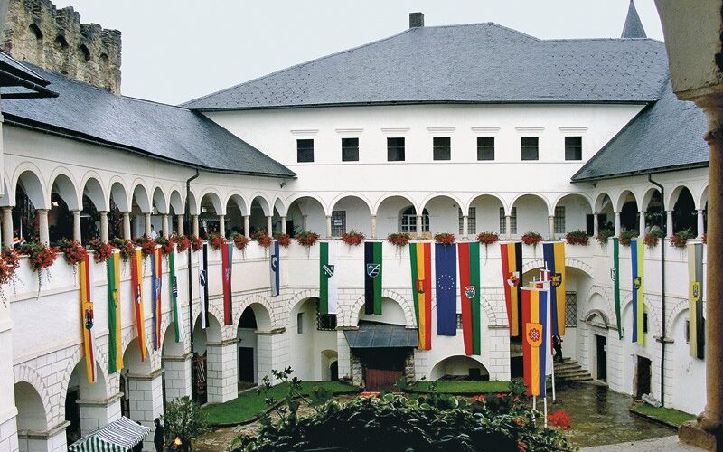 Ein Innenhof eines historischen Gebäudes mit gewölbten Gängen, roten Blumen und verschiedenen bunten vertikalen Fahnen, die von der oberen Ebene herabhängen.