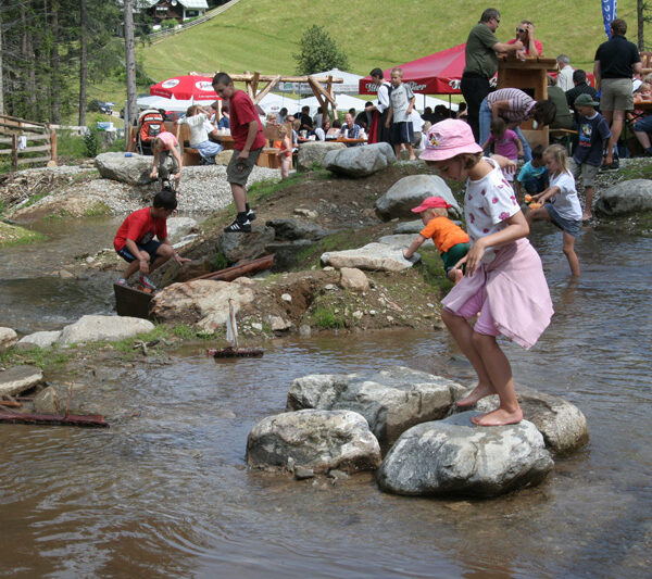Kinder und Erwachsene spielen und laufen auf Steinen in einem seichten Bach in einem Park, während im Hintergrund Menschen an Picknicktischen sitzen.