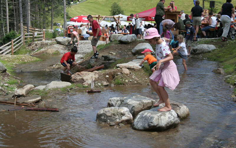 Kinder und Erwachsene spielen und laufen auf Steinen in einem seichten Bach in einem Park, während im Hintergrund Menschen an Picknicktischen sitzen.