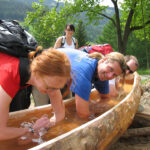 Vier Menschen lächeln, während sie ihre Arme in einem langen, ausgehöhlten Holztrog einweichen, der mit Wasser gefüllt ist, während im Hintergrund Bäume und Hügel zu sehen sind.