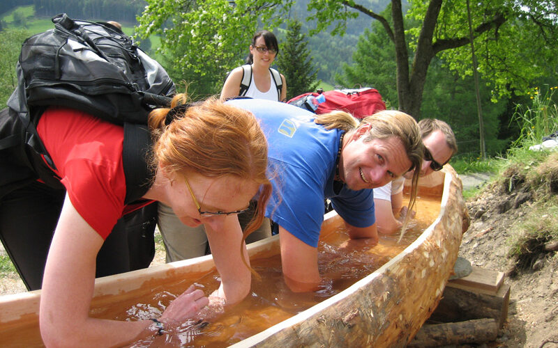 Vier Menschen lächeln, während sie ihre Arme in einem langen, ausgehöhlten Holztrog einweichen, der mit Wasser gefüllt ist, während im Hintergrund Bäume und Hügel zu sehen sind.
