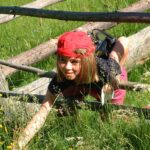 Ein kleines Kind mit roter Mütze und schwarzem Hemd krabbelt an einem sonnigen Tag unter einem Holzzaun hindurch und streckt sich durch hohes Gras und Wildblumen.