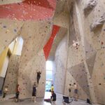 Indoor-Kletterhalle mit hohen Wänden, bunten Griffen, mehreren Kletterern und anderen, die beobachten oder am Boden warten.