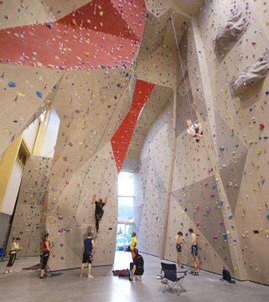 Indoor-Kletterhalle mit hohen Wänden, bunten Griffen, mehreren Kletterern und anderen, die beobachten oder am Boden warten.