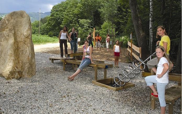 Eine Gruppe von Kindern und Erwachsenen steht und sitzt auf hölzernen Spielgeräten im Freien, umgeben von Bäumen und Kies.