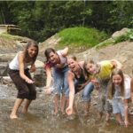 Sechs Kinder stehen in einem seichten Bach, spritzen Wasser und lächeln, mit einem Feldweg und grünen Bäumen im Hintergrund.