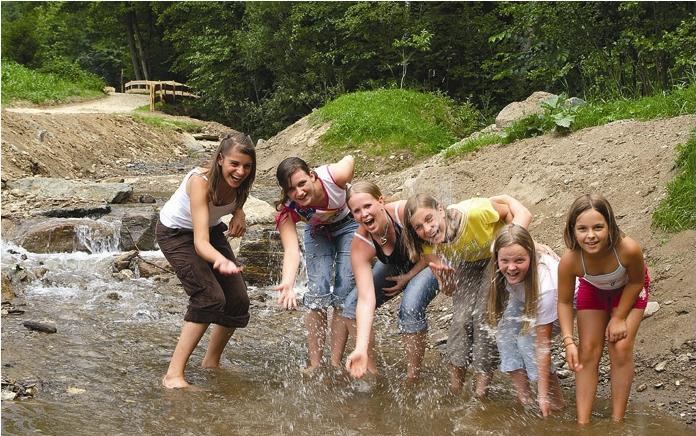 Sechs Kinder stehen in einem seichten Bach, spritzen Wasser und lächeln, mit einem Feldweg und grünen Bäumen im Hintergrund.