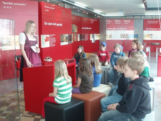 Eine Frau in traditioneller Kleidung spricht zu einer Gruppe von Kindern, die in einem Museum oder einer Ausstellung vor roten Informationsschildern sitzen.