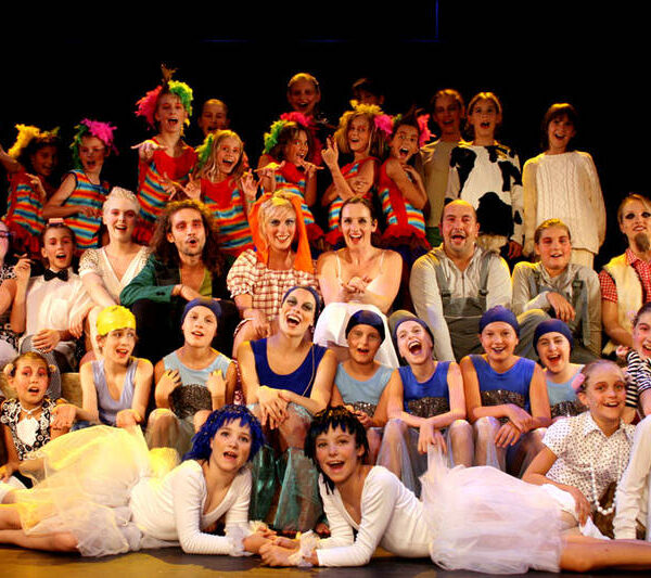 Eine große Gruppe von Kindern und Erwachsenen in farbenfrohen Kostümen posiert gemeinsam auf der Bühne und lächelt in die Kamera, wahrscheinlich im Rahmen einer Theater- oder Musicalaufführung.