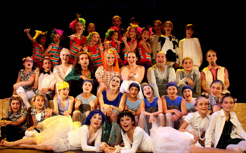 Eine große Gruppe von Kindern und Erwachsenen in farbenfrohen Kostümen posiert gemeinsam auf der Bühne und lächelt in die Kamera, wahrscheinlich im Rahmen einer Theater- oder Musicalaufführung.