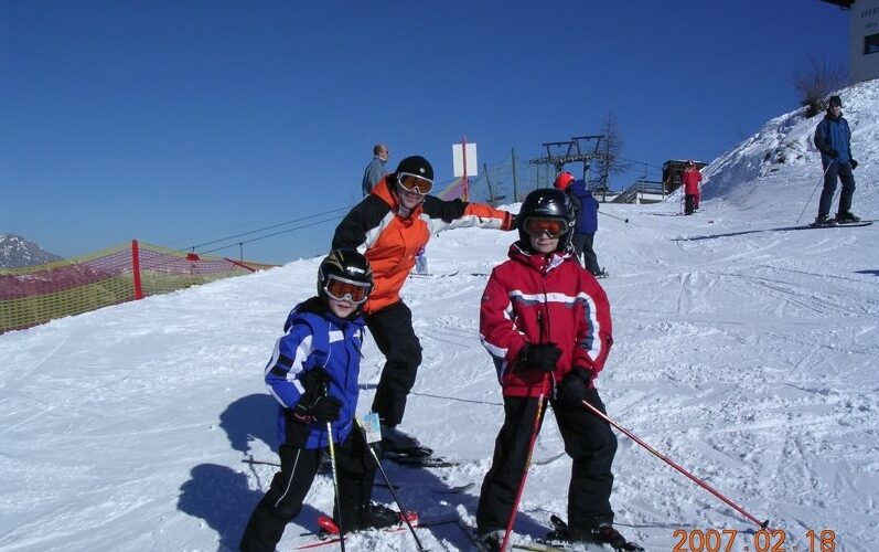 Drei Personen in Skiausrüstung posieren auf einer verschneiten Piste mit Skistöcken, während andere im Hintergrund zu sehen sind; das Datum lautet 2007-02-18.