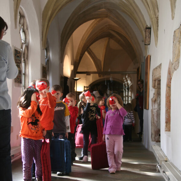 Eine Gruppe von Kindern mit Koffern geht in einer Reihe durch einen gewölbten Flur und hält sich rote Gegenstände vor das Gesicht.
