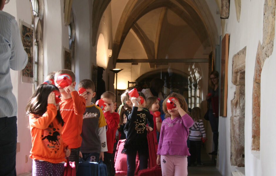 Eine Gruppe von Kindern mit Koffern geht in einer Reihe durch einen gewölbten Flur und hält sich rote Gegenstände vor das Gesicht.