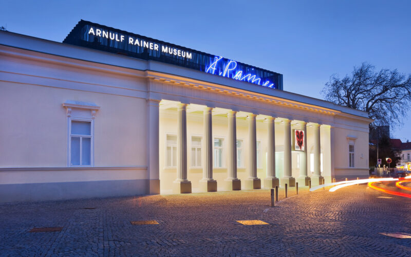 Das Gebäude des Arnulf-Rainer-Museums mit seinen neoklassizistischen Säulen wird in der Dämmerung beleuchtet, wobei die Lichtspuren der vorbeifahrenden Fahrzeuge im Vordergrund zu sehen sind.