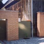 Zwei kleine Ziegel- und Schlackensteinbauten mit offenen Türen stehen neben einem Holzgebäude auf einer gepflasterten Fläche.