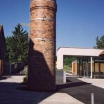 Ein zylindrischer Backsteinturm steht auf einer gepflasterten Fläche neben einem modernen Glasgebäude, mit einem klaren blauen Himmel im Hintergrund.