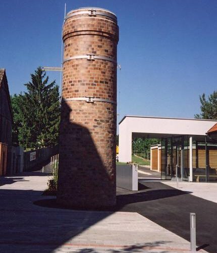 Ein zylindrischer Backsteinturm steht auf einer gepflasterten Fläche neben einem modernen Glasgebäude, mit einem klaren blauen Himmel im Hintergrund.