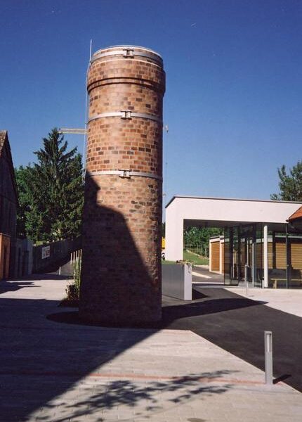 Ein zylindrischer Backsteinturm steht auf einer gepflasterten Fläche neben einem modernen Glasgebäude, mit einem klaren blauen Himmel im Hintergrund.
