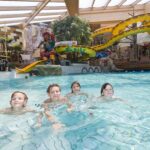 Vier Kinder schwimmen in einem Hallenbad mit einer Wasserrutsche im Piratenstil und einem Spielgerät im Hintergrund.