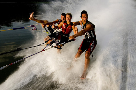 Drei Personen mit Schwimmwesten fahren nebeneinander auf einem See Wasserski, halten sich an Schleppseilen fest und das Wasser spritzt hinter ihnen auf.