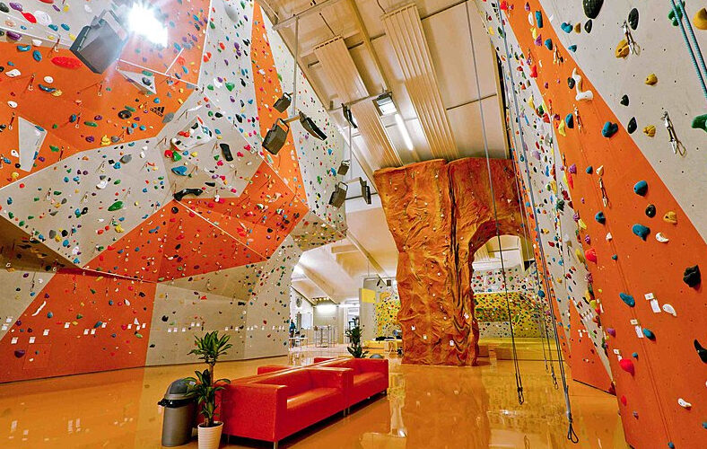 Indoor-Kletterhalle mit bunten Kletterwänden, einer künstlichen Felsstruktur, Seilen und einer roten Couch auf einem glänzenden Boden.
