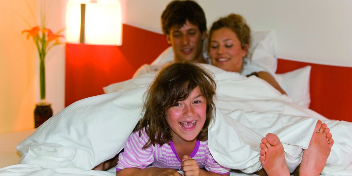 Ein lächelndes Kind liegt auf einem Bett vor zwei Erwachsenen unter einer weißen Decke in einem hell erleuchteten Schlafzimmer.