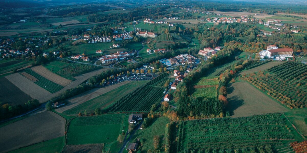 Luftaufnahme einer ländlichen Landschaft mit verstreuten Häusern, Ackerland, Obstgärten, kurvenreichen Straßen und entfernten Hügeln unter einem teilweise bewölkten Himmel.