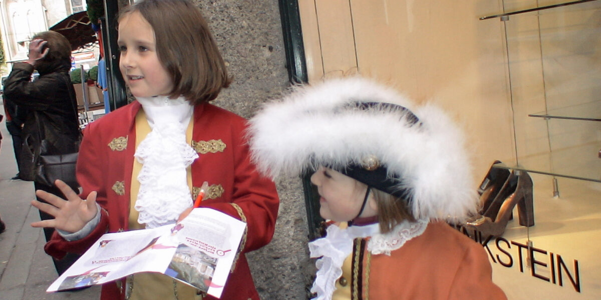 Zwei Kinder in historischen Kostümen stehen auf einem Bürgersteig; eines hält eine Broschüre in der Hand, während das andere einen großen Hut mit weißer Zierleiste trägt. Im Hintergrund ist die Auslage eines Schuhgeschäfts zu sehen.