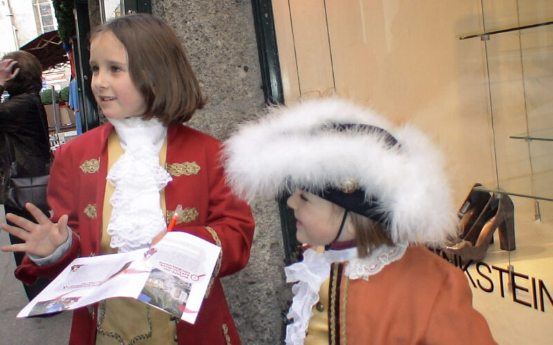 Zwei Kinder in historischen Kostümen stehen auf einem Bürgersteig; eines hält eine Broschüre in der Hand, während das andere einen großen Hut mit weißer Zierleiste trägt. Im Hintergrund ist die Auslage eines Schuhgeschäfts zu sehen.