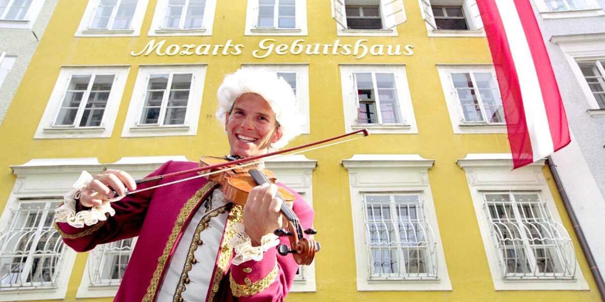 Eine Person in einem historischen Kostüm spielt Geige vor Mozarts Geburtshaus, einem gelben Gebäude mit der Aufschrift "Mozarts Geburtshaus" und einer österreichischen Flagge.
