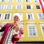 Eine Person in einem historischen Kostüm spielt Geige vor Mozarts Geburtshaus, einem gelben Gebäude mit der Aufschrift "Mozarts Geburtshaus" und einer österreichischen Flagge.
