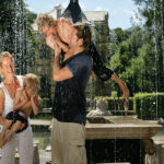 Zwei Erwachsene und zwei Kinder spielen an einem sonnigen Tag zusammen in einem Wasserbrunnen, wobei die Erwachsenen die Kinder hochheben, während das Wasser um sie herum herunterfließt.