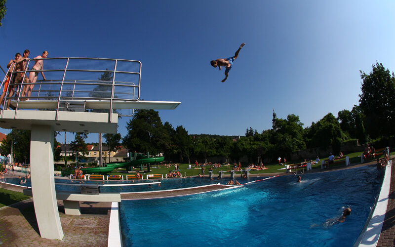 Eine Person befindet sich in der Luft, nachdem sie von einem Sprungbrett in ein Freibad gesprungen ist, während andere auf dem Sprungbrett warten und sich an einem sonnigen Tag um das Becken herum aufhalten.