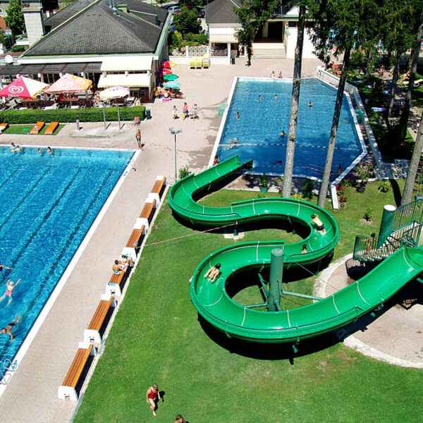 Luftaufnahme eines Freibadkomplexes mit zwei Becken, einer grünen Spiralwasserrutsche, Sonnenanbetern und schwimmenden Menschen.