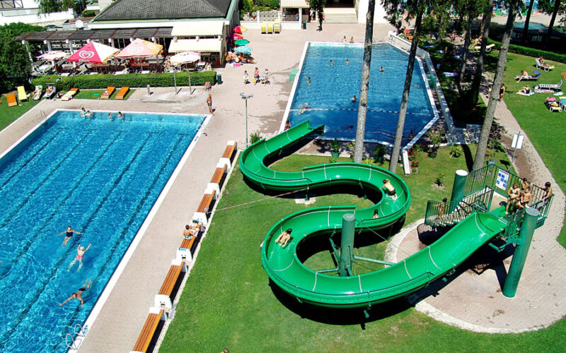 Luftaufnahme eines Freibadkomplexes mit zwei Becken, einer grünen Spiralwasserrutsche, Sonnenanbetern und schwimmenden Menschen.