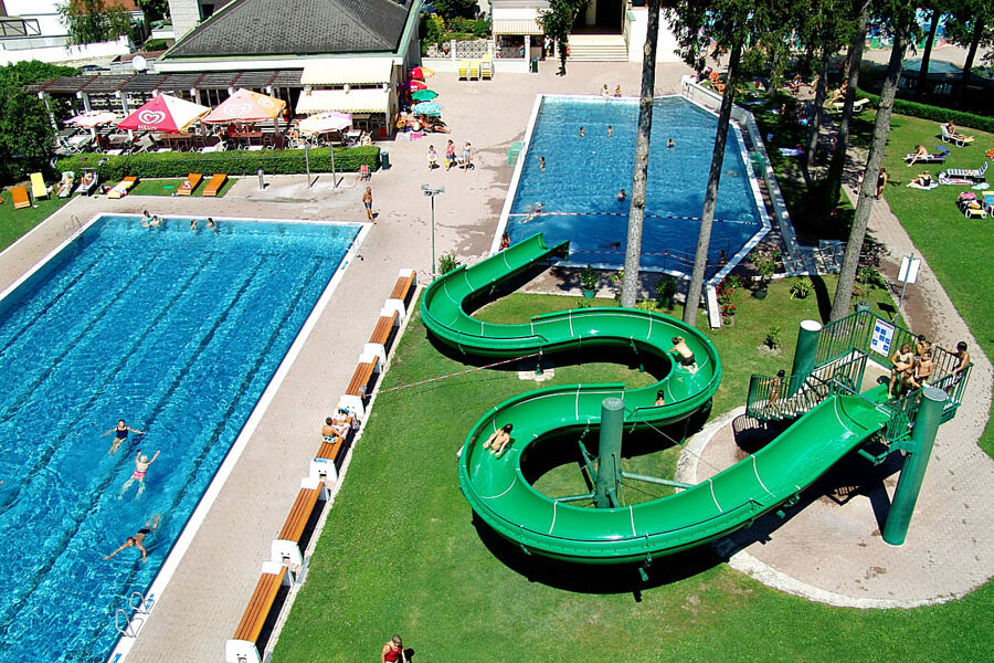 Luftaufnahme eines Freibadkomplexes mit zwei Becken, einer grünen Spiralwasserrutsche, Sonnenanbetern und schwimmenden Menschen.