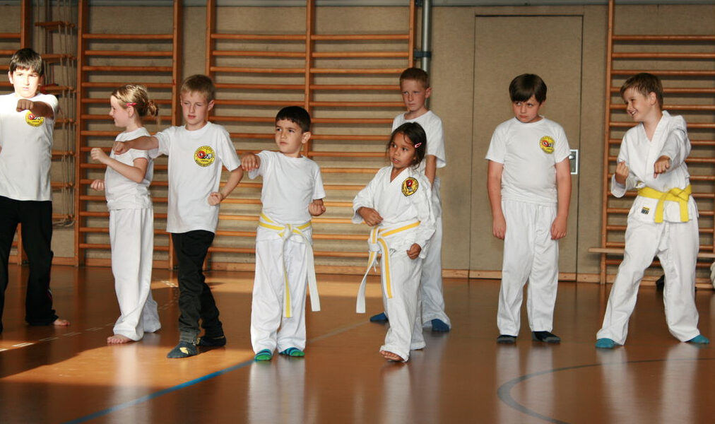 Eine Gruppe von Kindern in weißen Kampfsportuniformen und gelben Gürteln übt in einer Turnhalle mit hölzerner Sprossenwand Stellungen.