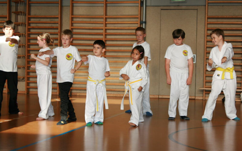 Eine Gruppe von Kindern in weißen Kampfsportuniformen und gelben Gürteln übt in einer Turnhalle mit hölzerner Sprossenwand Stellungen.